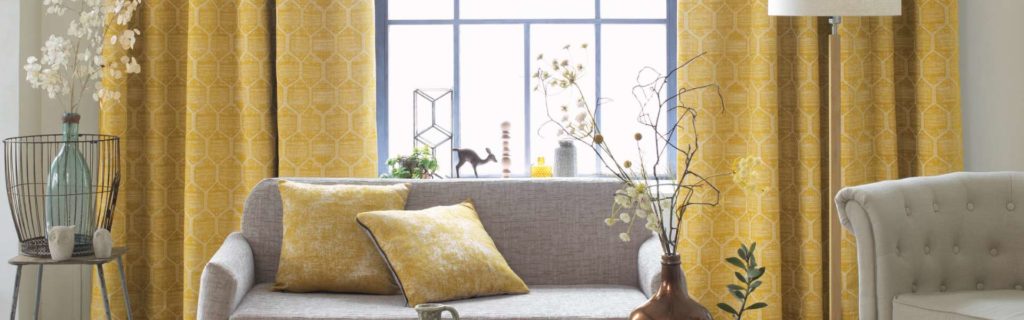 Bild Gardinen - Helles Wohnzimmer mit gelb gemusterten Vorhängen, einem grauen Sofa mit gelben Kissen und dekorativen Pflanzen neben einem großen Fenster. Raumausstattung Merget | Goethestraße 43 | 63814 Mainaschaff