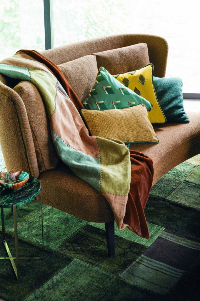 Ein gemütlicher Wohnraum mit einer braunen Couch, bedeckt mit bunten Decken und Kissen, neben einem Fenster mit Gardinen und Grün draußen. Raumausstattung Merget | Goethestraße 43 | 63814 Mainaschaff