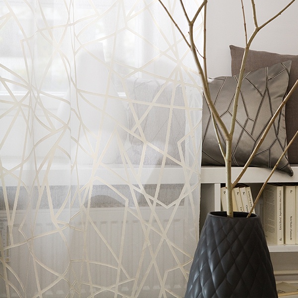 Transparente Gardinen mit geometrischem Muster in einem modernen Innenraumambiente, mit einer Vase mit Zweigen und einem Blick auf ein Bücherregal. Raumausstattung Merget | Goethestraße 43 | 63814 Mainaschaff