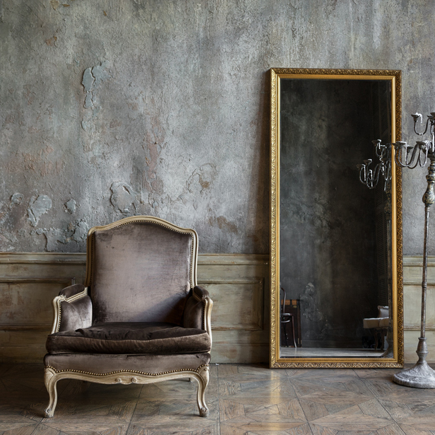 Ein antiker Sessel neben einem großen gerahmten Spiegel in einem Raum mit verwitterten Wänden und Gardinen. Raumausstattung Merget | Goethestraße 43 | 63814 Mainaschaff